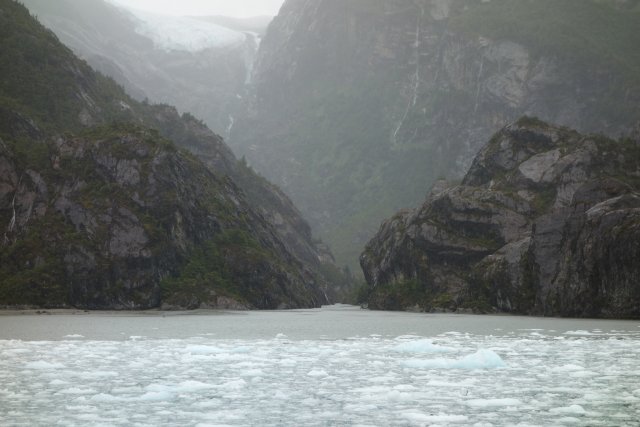 Schlucht am Rande des Fjordes Garibaldi. Die steilen Felswände steigen an den Seiten der Schlucht steil an. Das schmelzende Eis des weit oben gelegenen Gletschers bildet einen unaufhaltsamen Wasserstrom, der sich im Laufe der Jahrtausende durch das Gestein gegraben hat. Er scheint kein Eis mit sich zu führen, denn das Flussdelta ist eisfrei, während im Vordergrund eine dichte Schicht von Resten des Hauptgletschers vorbeizieht.