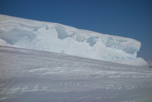 Bild der Schelfeiswand aus der Perspektive des Inlets, das sich bis zu zwanzig Meter in den Himmel streckt. Der Wind hat kunstvolle Skulpturen zum Vorschein gebracht, sodass dicke Wellen aus weißem Schnee über die Oberkante der Eiswand quillen wie Berge von Schlagsahne auf einem Stück Torte.